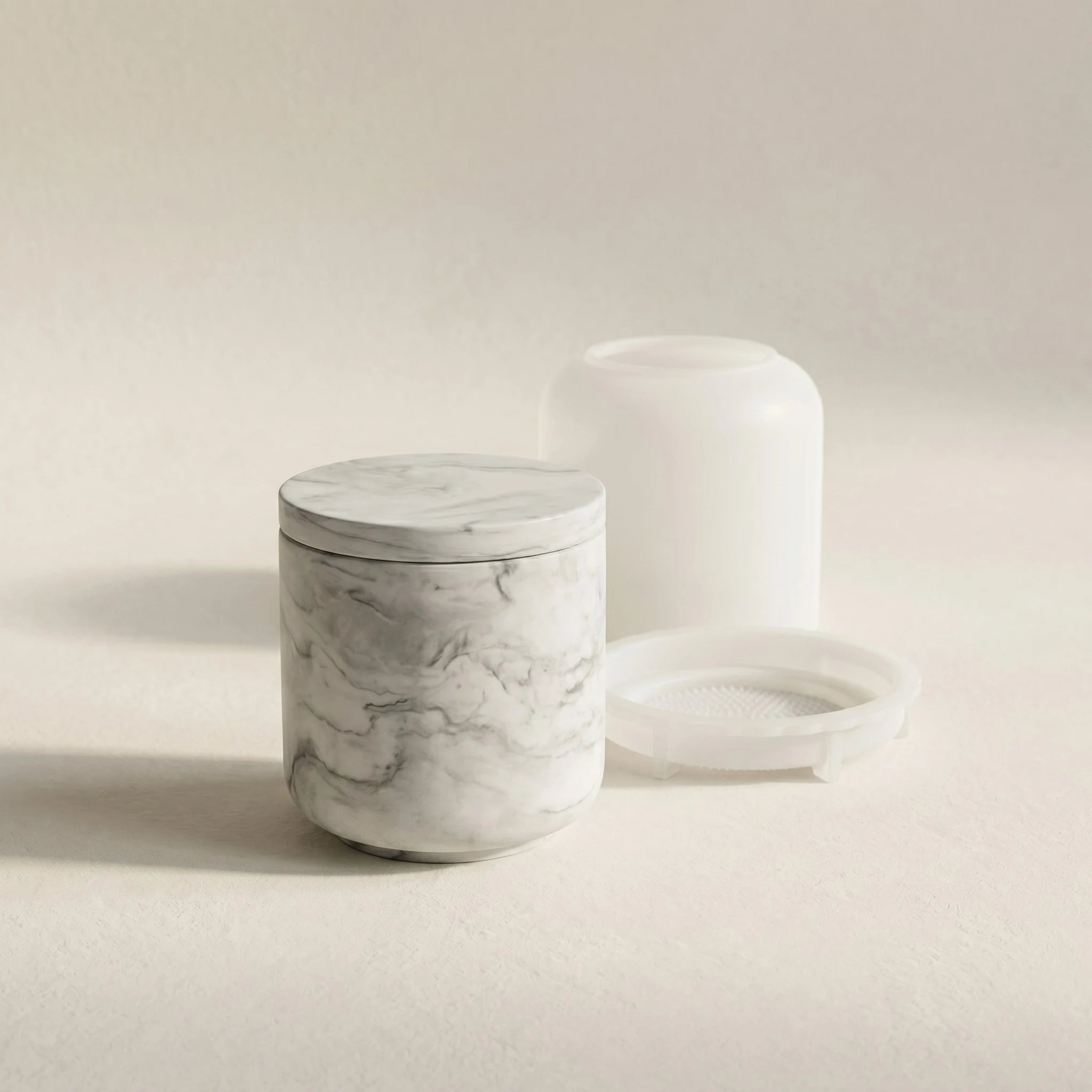 Minimalist grey marble cylindrical jar cast in eco-resin next to its translucent silicone mould set on a cream studio background.