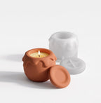 A small terracotta-colored candle pot with a lit wick and a matching lid placed in front, alongside a translucent white silicone mold behind it on a clean white background.